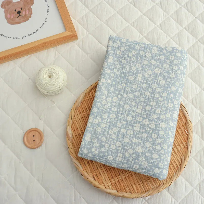 A folded yard of Longan Craft’s Small Floral Print Double Gauze Fabric—a botanical printed cotton crepe gauze material—rests on a wicker tray, accompanied by a spool of white thread, a large wooden button, and a framed bear illustration atop a quilted white surface.