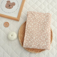A folded piece of Longan Craft’s Small Floral Print Double Gauze Fabric By The Yard rests on a round wicker tray, accompanied by a ball of white yarn, a wooden button, and a framed bear illustration arranged on a quilted white surface.