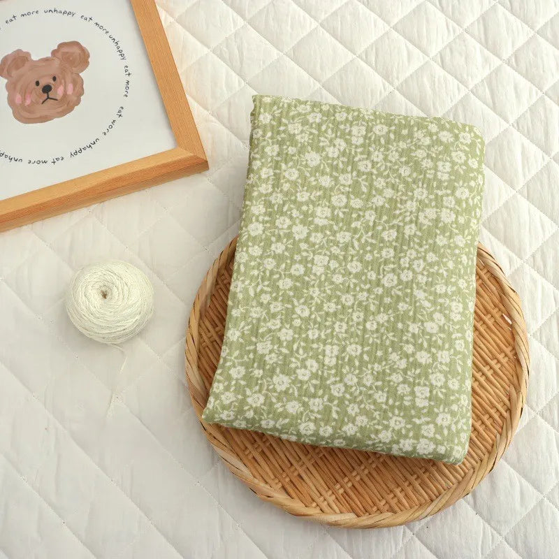 A piece of Longan Craft’s Small Floral Print Double Gauze Fabric By The Yard, featuring a green and white botanical design on soft cotton crepe gauze, is neatly folded on a round wicker tray. Next to it sit a ball of white yarn and a framed bear illustration, all arranged atop a quilted white surface.