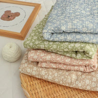 Four pieces of the Small Floral Print Double Gauze Fabric By The Yard from Longan Craft, featuring gentle botanical prints in pastel shades, are folded and stacked atop a woven basket. Beside them on a white surface sit a ball of white yarn and a framed illustration of a bear.