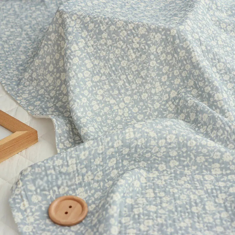 A piece of Longan Craft’s Small Floral Print Double Gauze Fabric By The Yard—a light blue, botanical-printed cotton crepe gauze material—is draped loosely, showing its gentle texture. In the bottom left corner lies a brown button, while a wooden frame is placed next to the fabric on a white surface.