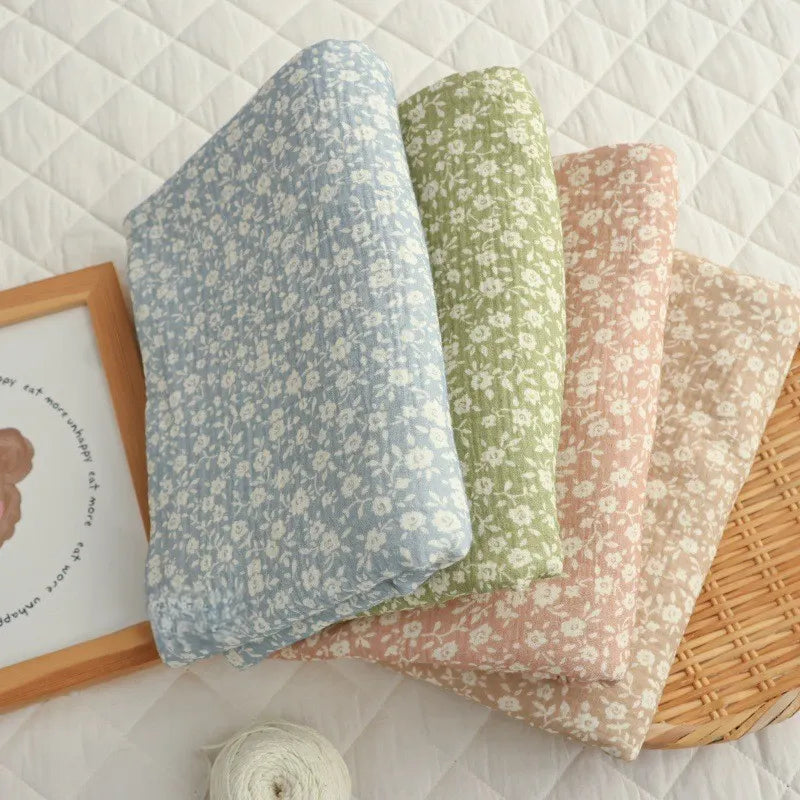 Four folded pieces of Longan Craft’s Small Floral Print Double Gauze Fabric, in blue, green, pink, and beige—each crafted from soft botanical printed cotton crepe gauze—are neatly displayed in a row on a quilted white surface beside a framed picture and a ball of white yarn.