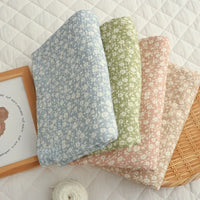 Four folded pieces of Longan Craft’s Small Floral Print Double Gauze Fabric, in blue, green, pink, and beige—each crafted from soft botanical printed cotton crepe gauze—are neatly displayed in a row on a quilted white surface beside a framed picture and a ball of white yarn.