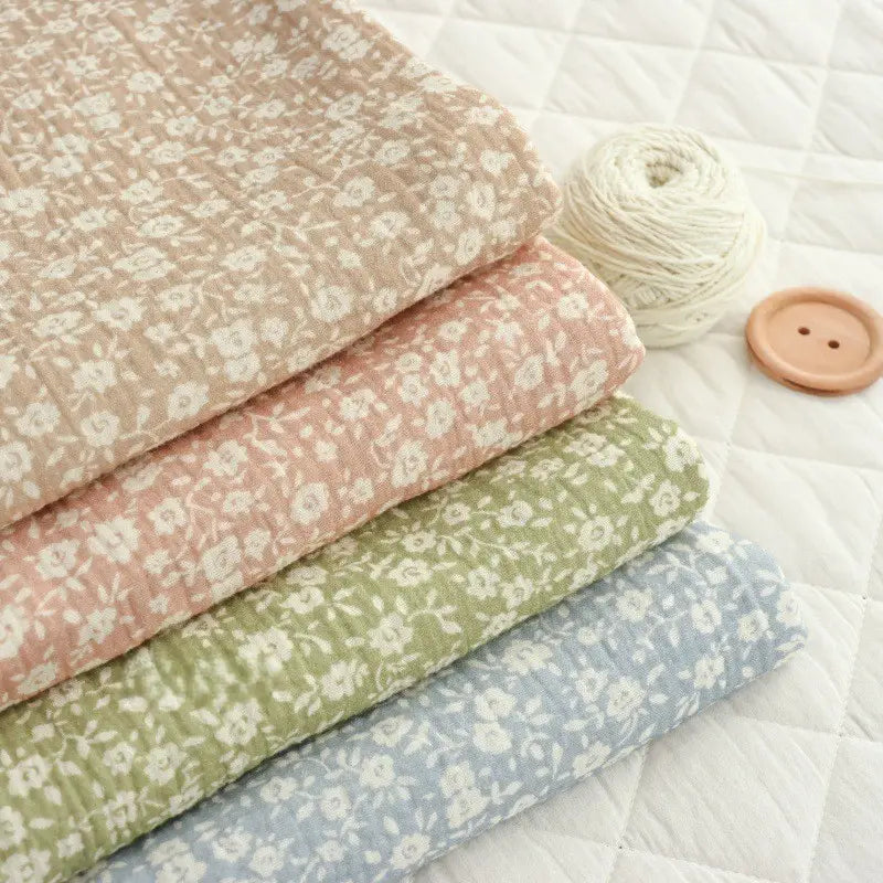 Four folded pieces of Longan Craft Small Floral Print Double Gauze Fabric By The Yard in beige, pink, green, and blue are displayed on a white quilted surface beside a ball of cream yarn and a large wooden button.
