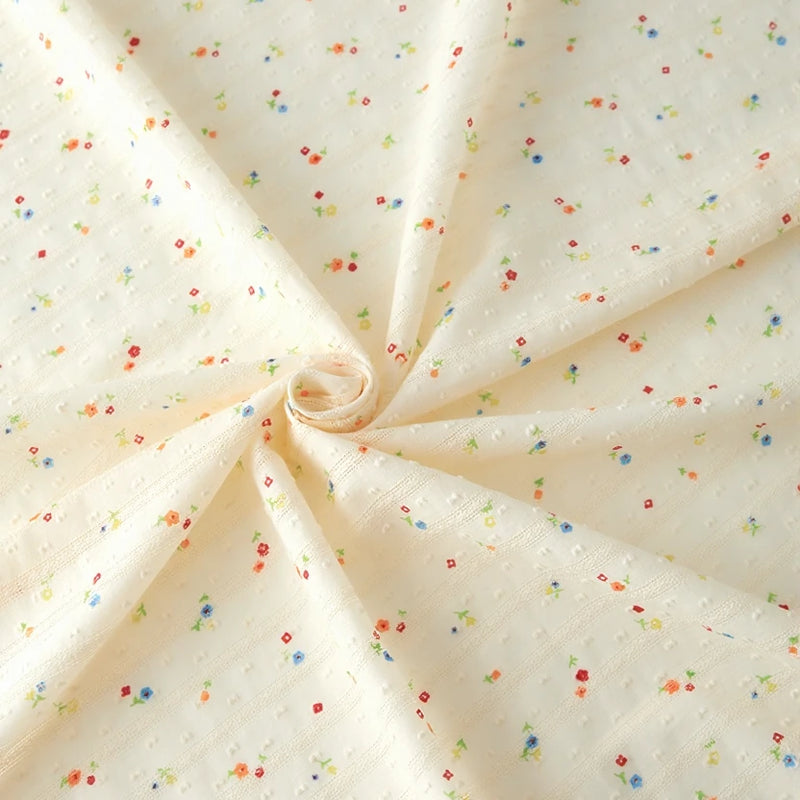 Small Floral Printed Swiss Dot Fabric by Longan Craft features cozy cream cotton with a dotted texture, accented by tiny red, blue, green, and yellow floral prints. Perfect for clothing; fabric shown elegantly swirled.