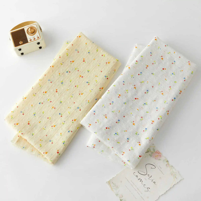 Two pieces of Small Floral Printed Swiss Dot Fabric by Longan Craft, one cream and one white with colorful flowers, are displayed on a white surface next to a vintage radio and a floral-patterned card.