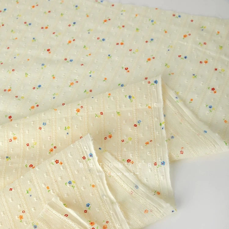 Longan Craft’s Small Floral Printed Swiss Dot Cotton Fabric features thin textured stripes and scattered red, blue, green, and orange flowers on cream, ideal for clothing. Sold by the yard; some edges are shown folded over.