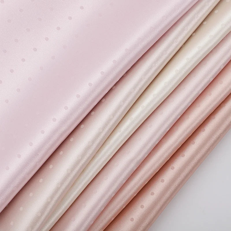 A close-up of neatly stacked Longan Craft Small Polka Dot Jacquard Satin Fabric By The Yard, showing pastel pink and cream shades with a subtle multicolor dotted jacquard satin texture, perfect for cozy clothing.