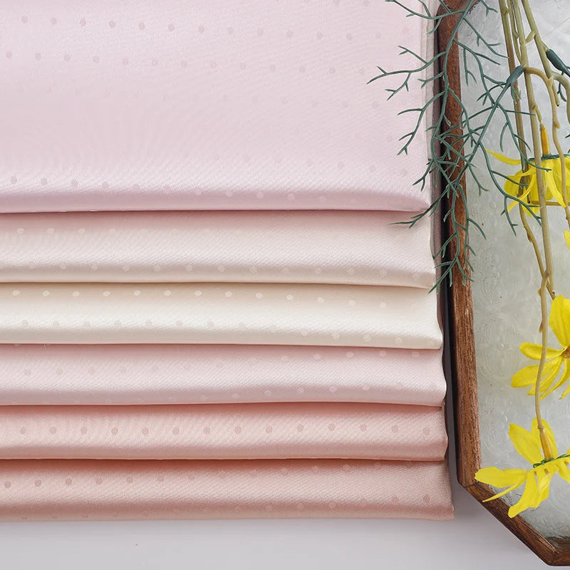 Small Polka Dot Jacquard Satin Fabric by the yard from Longan Craft, in pastel pink and cream, is neatly stacked beside yellow flowers and green leaves, with a hint of a wooden tray on the right. Perfect cozy satin material for clothing.