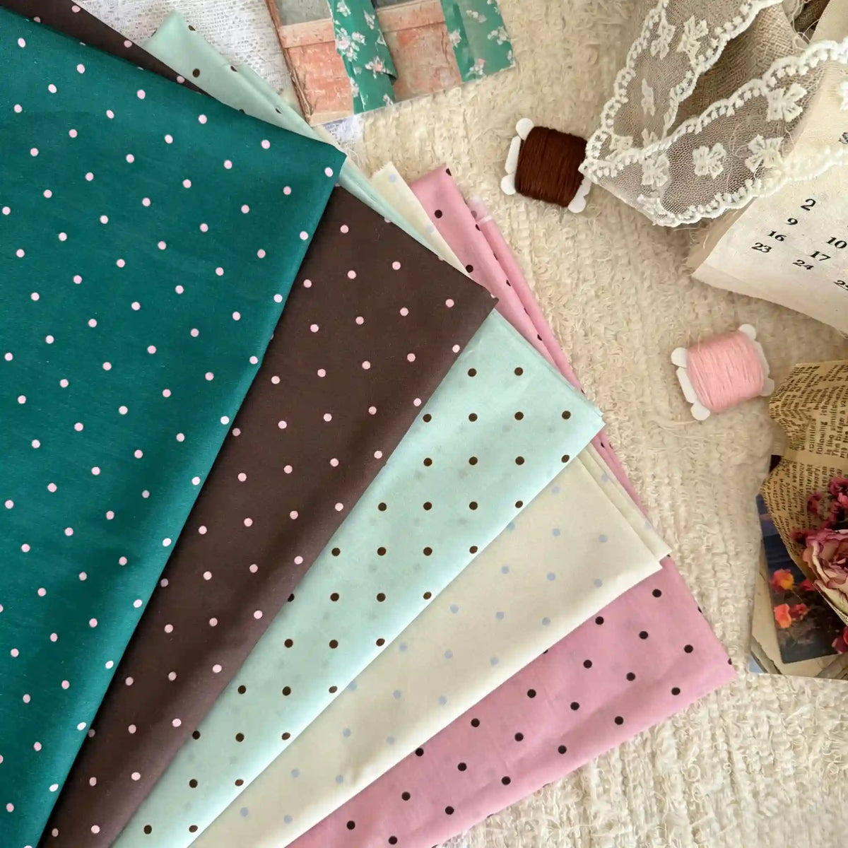Five pieces of Longan Craft’s Small Polka Dot Printed 100% Cotton Lawn Fabric in teal, brown, mint, cream, and pink are displayed on a textured surface with lace, thread spools, a calendar, and floral-patterned paper nearby.