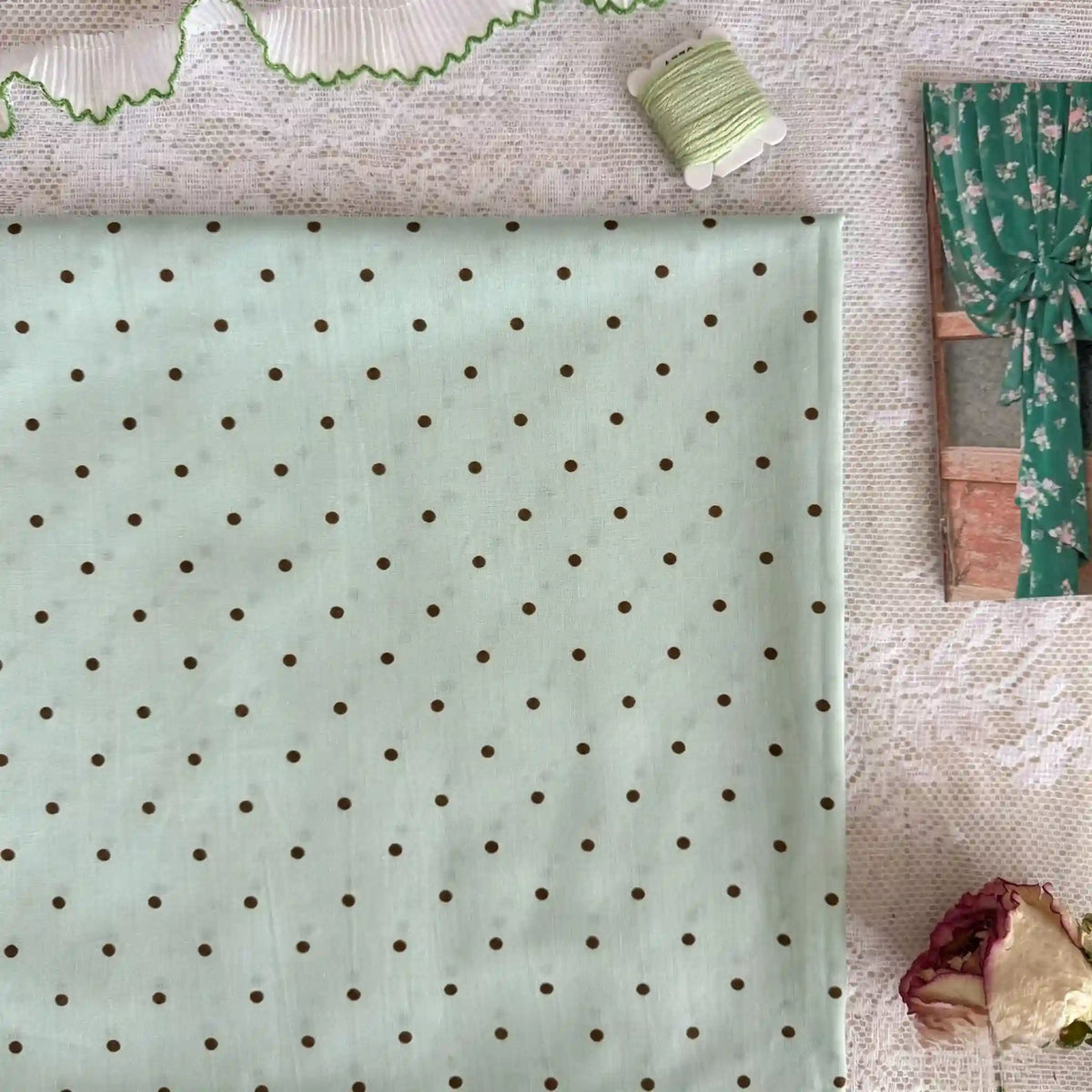 A folded Longan Craft Small Polka Dot Printed Lawn Fabric (100% cotton, pastel green) rests on lace, with green thread, ruffled trim, a floral curtain photo, and a dried rose nearby.