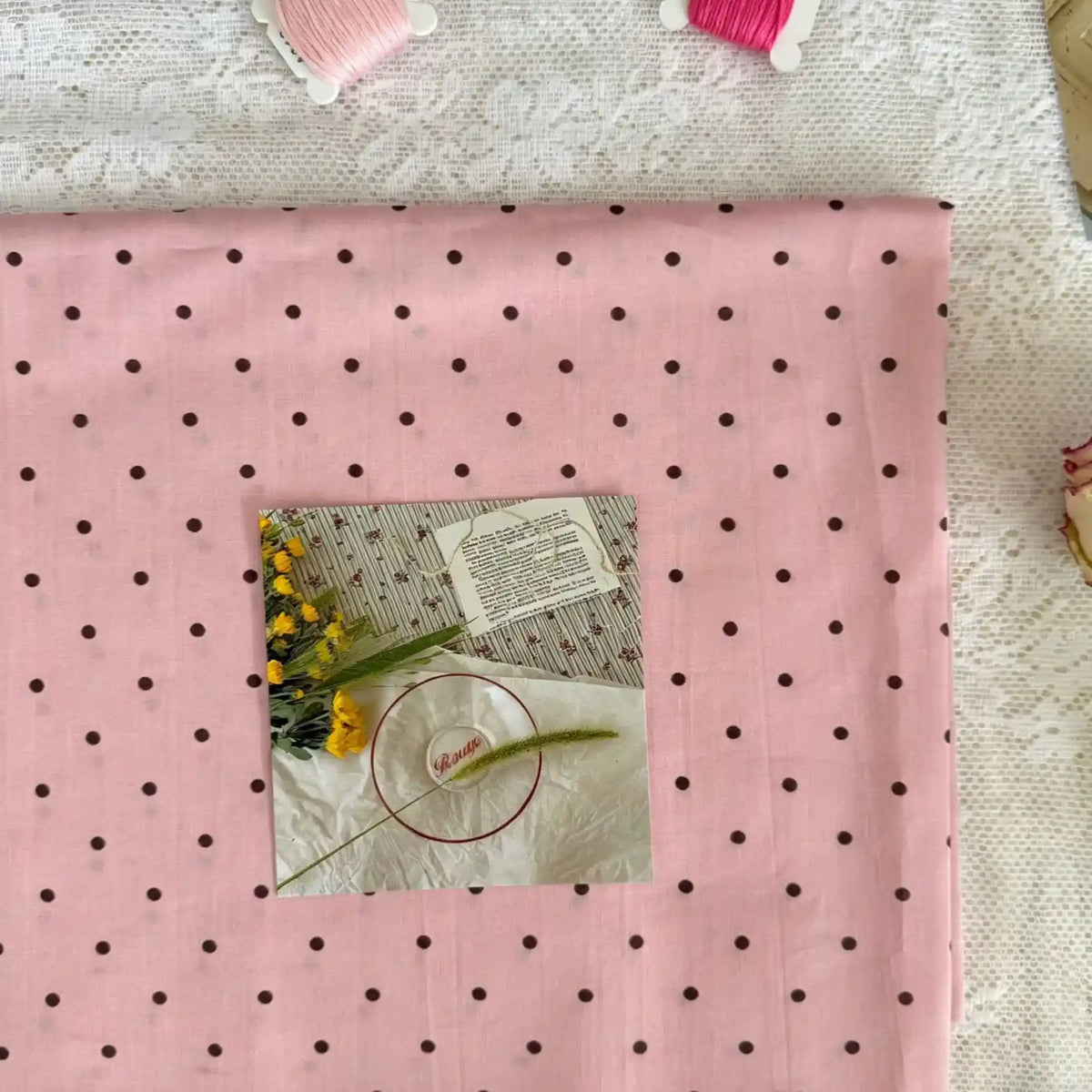 A folded piece of Small Polka Dot Printed Lawn Fabric by Longan Craft rests on lace. On top are embroidery supplies, yellow flowers, and sewing instructions, with pink thread spools nearby. This lightweight 100% cotton fabric is ideal for casual garments.