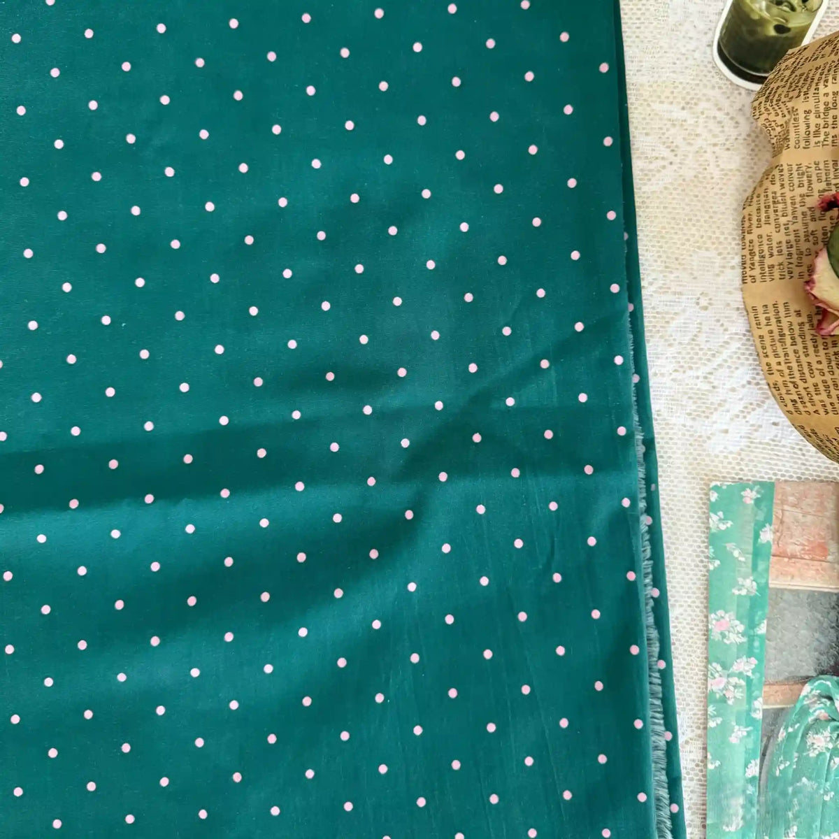 A teal Small Polka Dot Printed Lawn Fabric by Longan Craft, made of lightweight 100% cotton, lies on lace beside patterned paper, a woven basket with dried flowers, and a container—perfect materials for creative projects.