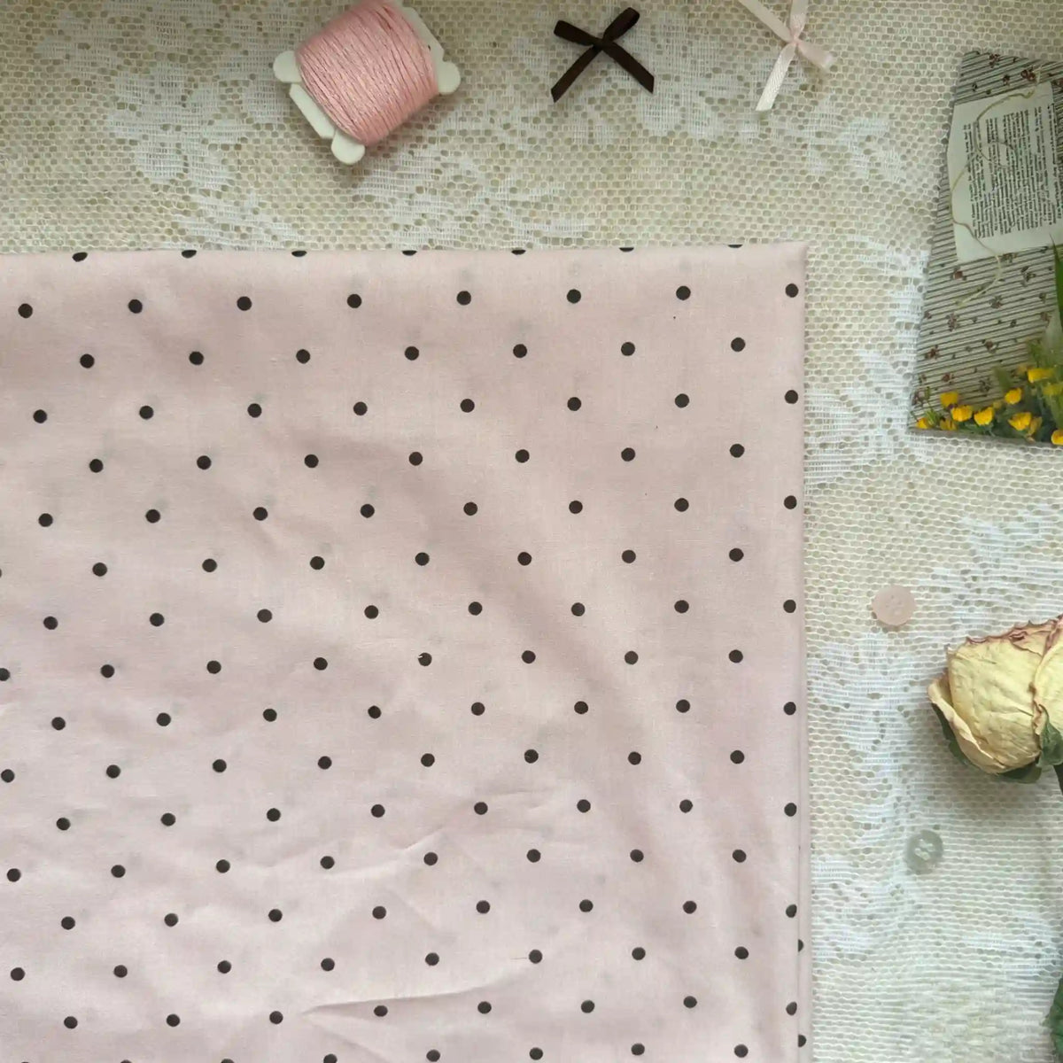 A piece of Longan Craft Small Polka Dot Printed Lawn Fabric By The Yard, a lightweight 100% cotton fabric for casual garments, rests on lace with pink thread, a ribbon, a dried rose, and a floral packet.