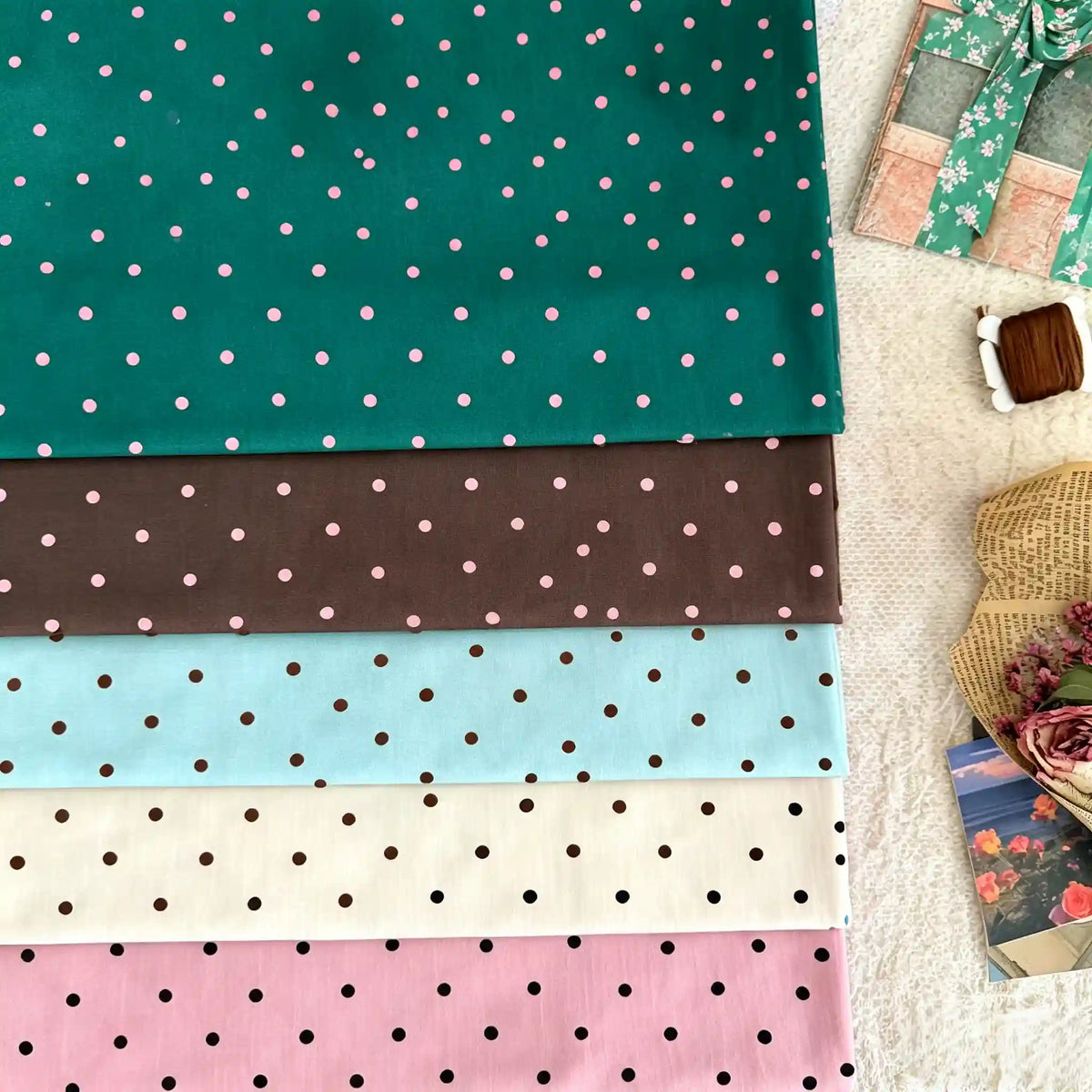 Folded Longan Craft Small Polka Dot Printed Lawn Fabric by the yard in green, brown, light blue, cream, and pink rests on a textured surface with a wrapped gift, thread spool, woven basket, and dried flowers nearby.
