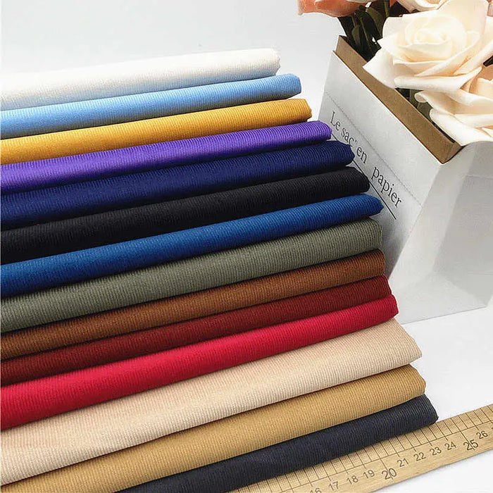 Solid Color 21 Wales Corduroy Fabric by the Yard from Longan Craft is displayed in red, blue, yellow, beige, and brown rolls beside a box of artificial white roses and a wooden ruler in the foreground.