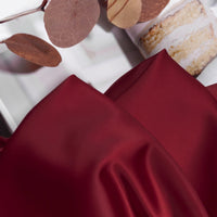 A close-up of Longan Craft Solid Color Charmeuse Satin Fabric By The Yard with a thick bridal satin texture, shown in deep red next to dried eucalyptus leaves and a slice of layered cake on a white plate.