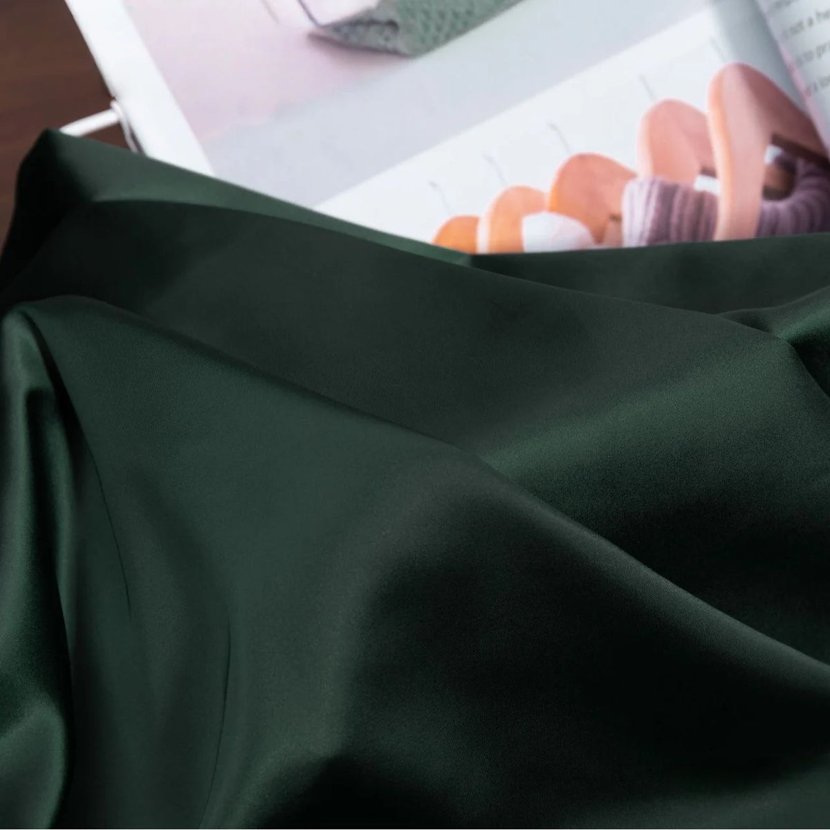 A close-up of Longan Craft Solid Color Charmeuse Satin Fabric by the Yard—thick bridal satin with a smooth, shiny finish, draped in soft folds; an open magazine featuring wooden hangers and clothing appears in the background.