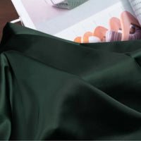A close-up of Longan Craft Solid Color Charmeuse Satin Fabric by the Yard—thick bridal satin with a smooth, shiny finish, draped in soft folds; an open magazine featuring wooden hangers and clothing appears in the background.