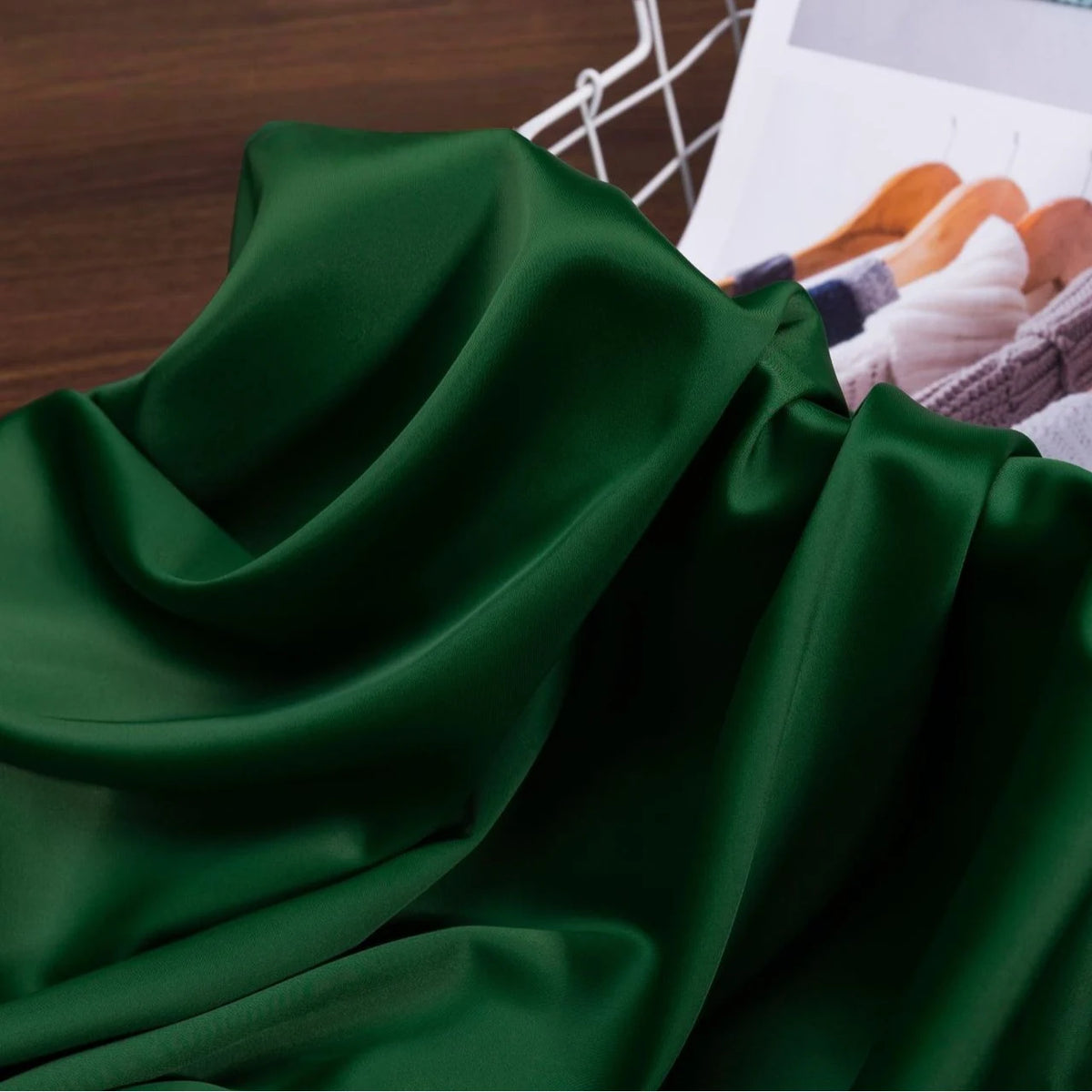 A Solid Color Charmeuse Satin Fabric by the yard from Longan Craft, with a thick bridal satin texture, is draped on wood beside a magazine featuring sweaters on hangers and a white wire basket.