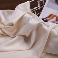 Close-up of Longan Craft’s Solid Color Charmeuse Satin Fabric By The Yard, featuring a thick bridal satin texture and smooth, luxurious finish—perfect for elegant gowns; blurred magazine and white wire rack in the background.