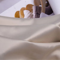 A close-up of Longan Craft's Solid Color Charmeuse Satin Fabric by the yard, featuring its thick bridal satin texture and smooth finish, with blurred wooden hangers and a rolled-up magazine in the background.