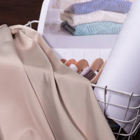 A Solid Color Charmeuse Satin Fabric by the yard from Longan Craft is draped over a white wire basket holding a rolled sheet and a printed image of folded pastel knit sweaters on hangers.