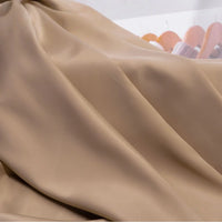 Smooth, light brown Solid Color Charmeuse Satin Fabric by the yard from Longan Craft drapes in gentle folds, showcasing its thick bridal satin texture; blurred wooden hangers and faint clothing appear in the background.
