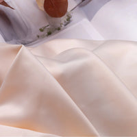 A close-up of Longan Craft Solid Color Charmeuse Satin Fabric By The Yard, showcasing its smooth, thick bridal satin texture in light beige with soft folds; a blurred figure in a white dress and brown accessory is visible in the background.