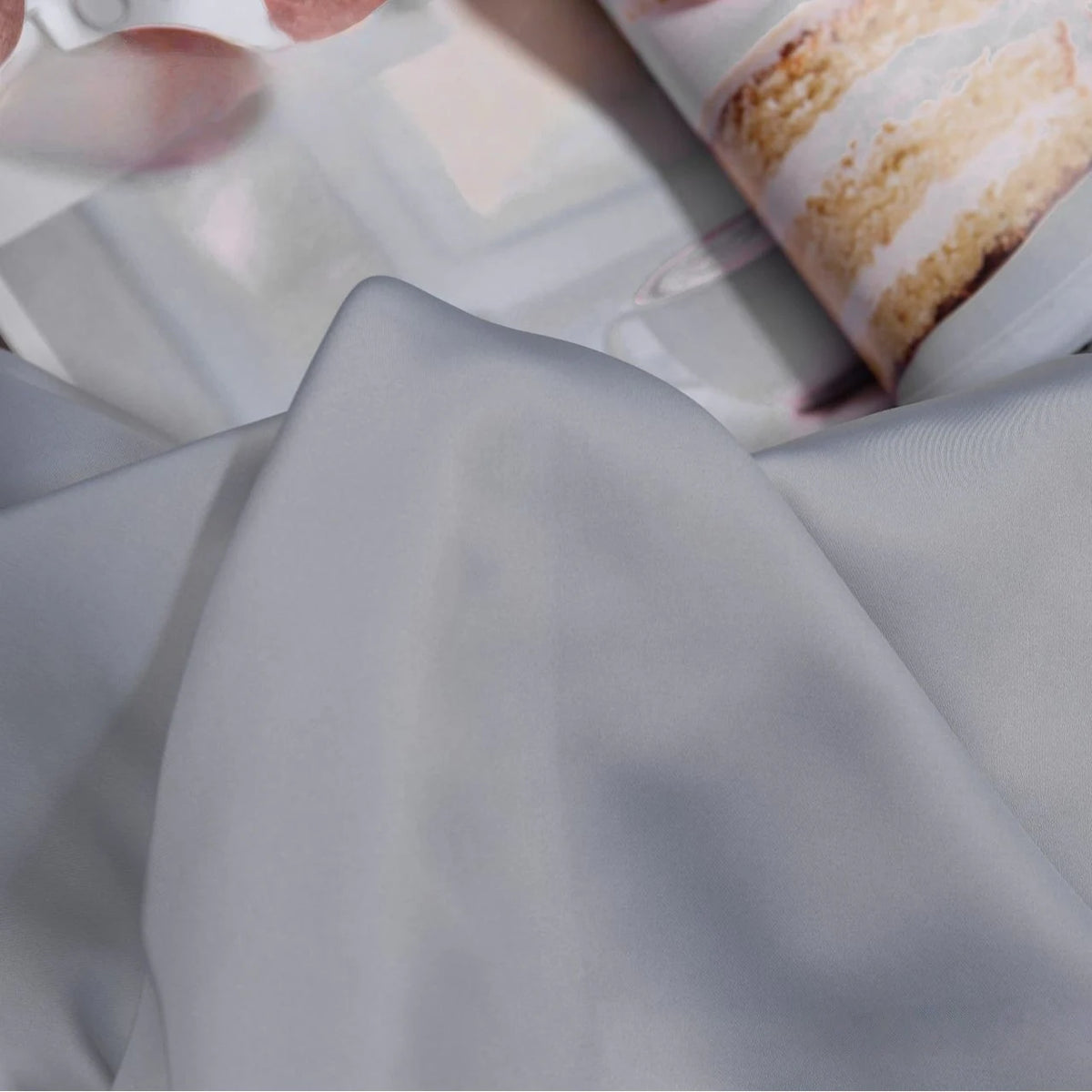 A close-up shows Longan Craft Solid Color Charmeuse Satin Fabric By The Yard, thick bridal satin with a smooth texture, in light gray, draped over a magazine page featuring a slice of cake with white icing.