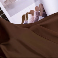 A Solid Color Charmeuse Satin Fabric by Longan Craft is draped in the foreground, its smooth, thick bridal satin texture displayed, with an open magazine showing clothing on wooden hangers in the background.