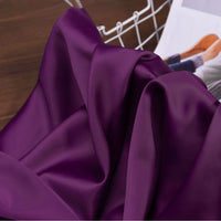 A piece of Longan Craft Solid Color Charmeuse Satin Fabric by the yard, featuring thick bridal satin texture, is draped on wood with part of a white wire basket and a magazine or book in the background.