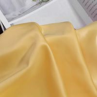 A close-up of Longan Craft Solid Color Charmeuse Satin Fabric By The Yard, featuring thick bridal satin texture in light yellow, elegantly draped with a partially visible open magazine in the background.