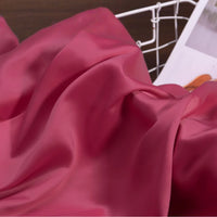 A close-up of Longan Craft’s Solid Color Charmeuse Satin Fabric By The Yard, featuring thick bridal satin texture in smooth pink, draped over a surface with a white wire basket and magazine in the background.
