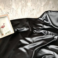 A yard of Longan Craft's Solid Color Charmeuse Satin Fabric, a thick bridal satin with a smooth texture, is draped across a surface. In the upper left, a flamingo art print rests against a textured light wall.