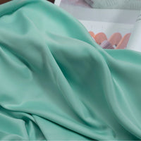 A close-up of Longan Craft Solid Color Charmeuse Satin Fabric By The Yard, featuring thick bridal satin texture in mint green, draped in soft folds with pastel clothes on hangers softly blurred in the background.
