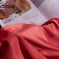 A smooth, coral-pink Longan Craft Solid Color Charmeuse Satin Fabric is draped in the foreground, with dried brown leaves resting on an open magazine in the background.