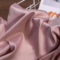 A close-up of Longan Craft Solid Color Charmeuse Satin Fabric By The Yard, featuring its thick, smooth, and shiny bridal satin texture—ideal for luxury gown projects. A wire basket and a blurred magazine appear in the background.