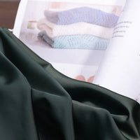A close-up of Longan Craft Solid Color Charmeuse Satin Fabric By The Yard, with its thick bridal satin texture, is draped over an open magazine showing folded knit blankets in white, blue, and gray.