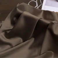 A close-up of Longan Craft's Solid Color Charmeuse Satin Fabric By The Yard, featuring a thick bridal satin texture with smooth, shiny brown folds on a dark surface. White wires and a partially visible paper appear in the background.