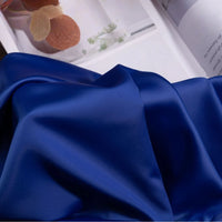 A smooth, shiny royal blue Solid Color Charmeuse Satin Fabric by Longan Craft is draped in the foreground, with part of an open magazine showing plant images and text in the background.