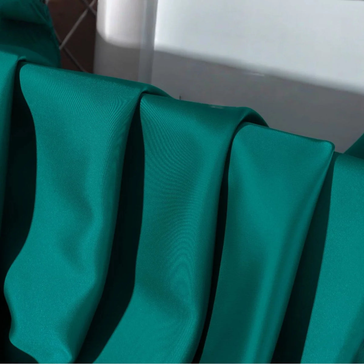 Close-up of Longan Craft Solid Color Charmeuse Satin Fabric By The Yard, featuring thick bridal satin texture in teal-green, draped in smooth folds with soft lighting showing its sheen. A white background surface is visible.