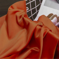 A close-up of Longan Craft Solid Color Charmeuse Satin Fabric by the yard, featuring its thick bridal satin texture in vibrant orange, elegantly draped in soft folds with a magazine and white grid in the background.