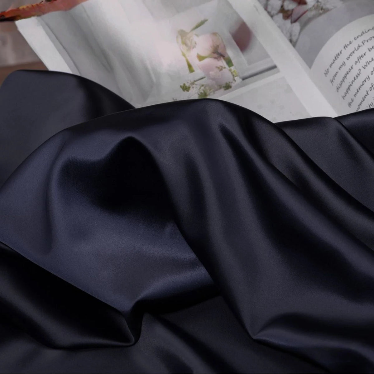 A close-up of Longan Craft Solid Color Charmeuse Satin Fabric By The Yard, featuring thick bridal satin with a smooth texture, draped in soft folds and an open magazine with photos and text in the background.