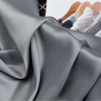 A close-up of Longan Craft’s Solid Color Charmeuse Satin Fabric By The Yard reveals its thick, smooth bridal satin texture. In the background, a blurred magazine on hangers highlights this elegant textile for gowns.