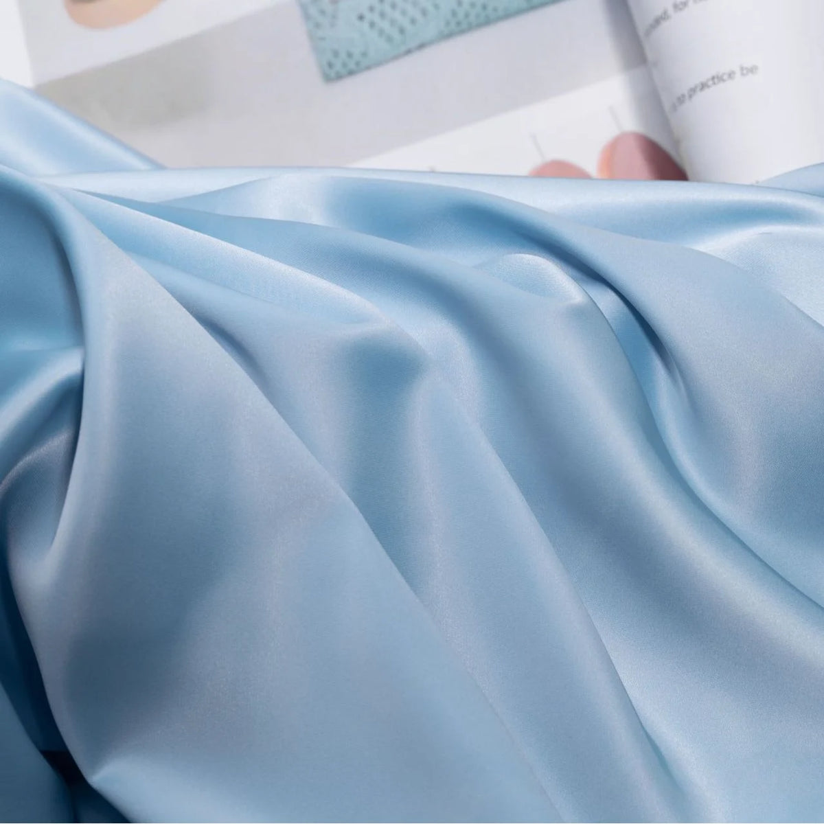 Longan Craft’s Solid Color Charmeuse Satin Fabric By The Yard is draped in soft folds over an open magazine, showcasing its thick bridal satin texture and smooth, silky elegance—ideal for luxurious gowns.