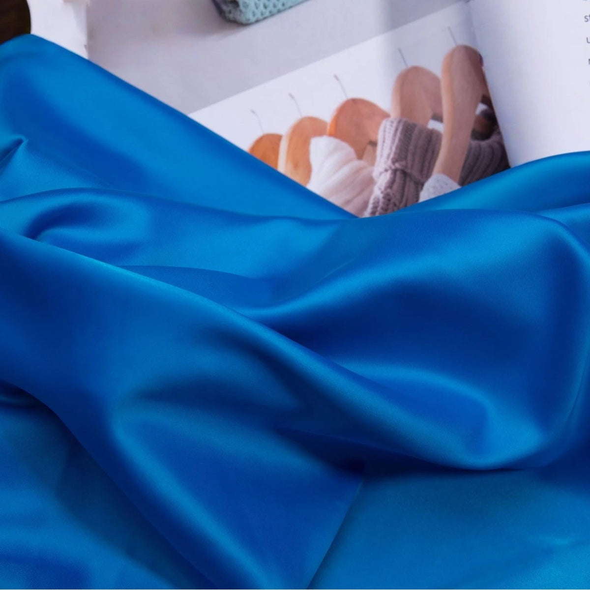 The Longan Craft Solid Color Charmeuse Satin Fabric By The Yard, a thick bridal satin with a smooth finish, is displayed in blue with soft folds, while an open magazine in the background features wooden hangers and knitwear.