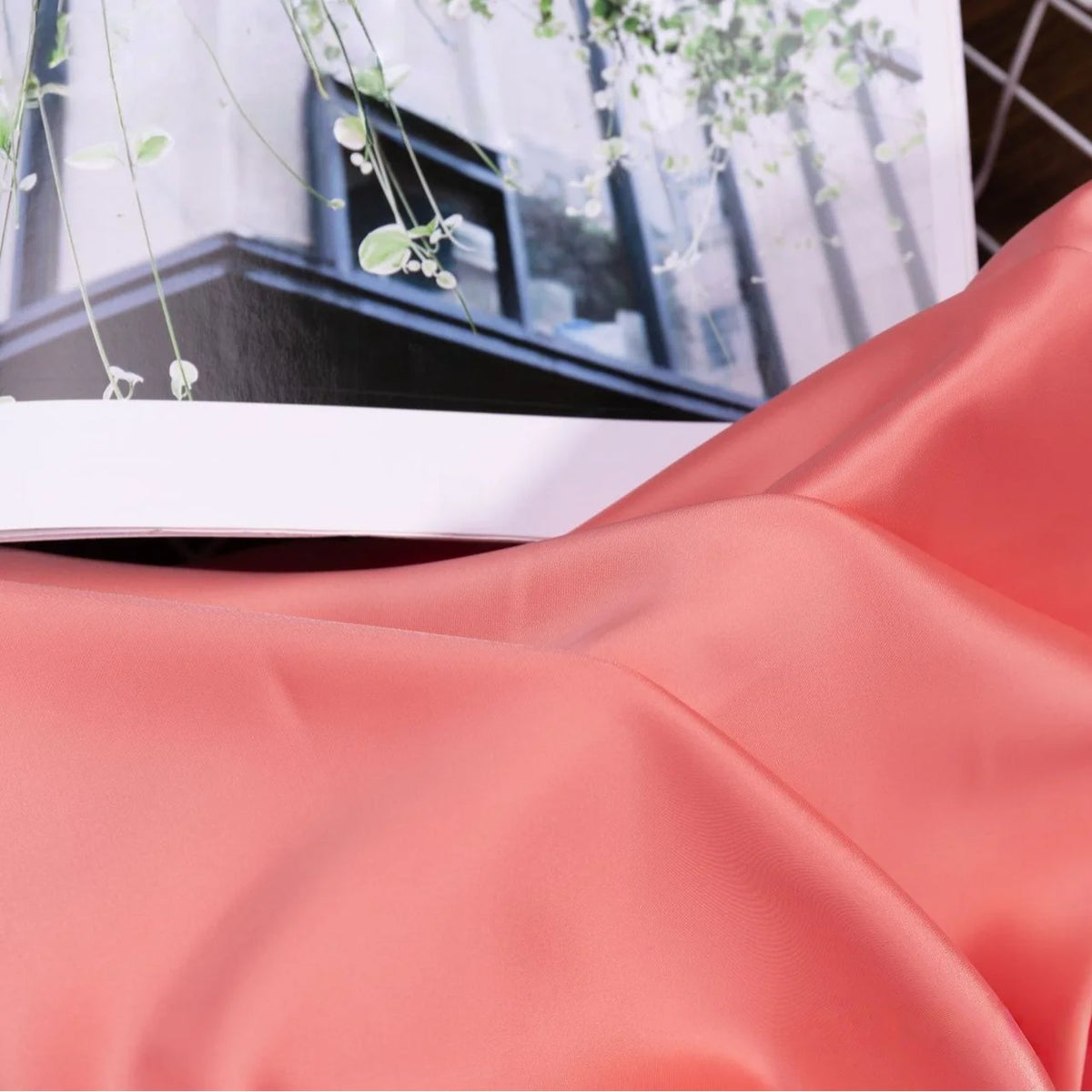 A smooth, peach-colored Solid Color Charmeuse Satin Fabric By The Yard from Longan Craft is draped in the foreground, with an open magazine displaying green hanging plants and a building in the background.