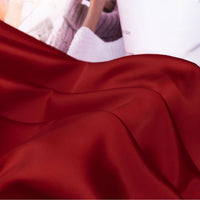 A close-up of Longan Craft Solid Color Charmeuse Satin Fabric By The Yard showcases its smooth, shiny red bridal satin texture with gentle folds; a white cup and a person in a cozy sweater are blurred in the background.