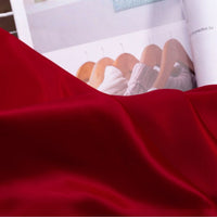A close-up of Longan Craft’s Solid Color Charmeuse Satin Fabric By The Yard, featuring vibrant red thick bridal satin texture in the foreground, with an open magazine in the background displaying sweaters on hangers.